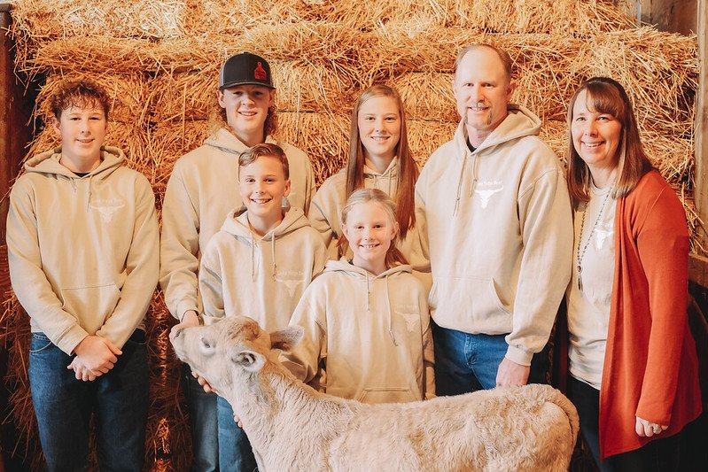 Family portrait in a barn setting, featuring six members and a calf, all dressed in matching hoodies. The backdrop consists of stacked hay bales, highlighting a farm environment.
