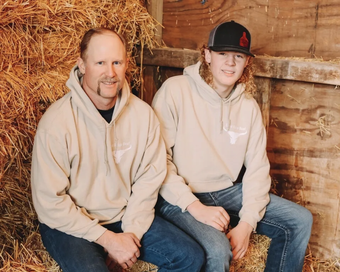 Father and son duo sitting on a haystack in a barn, both wearing beige hoodies with a beef brand logo, showcasing a rustic farming lifestyle and family bond.