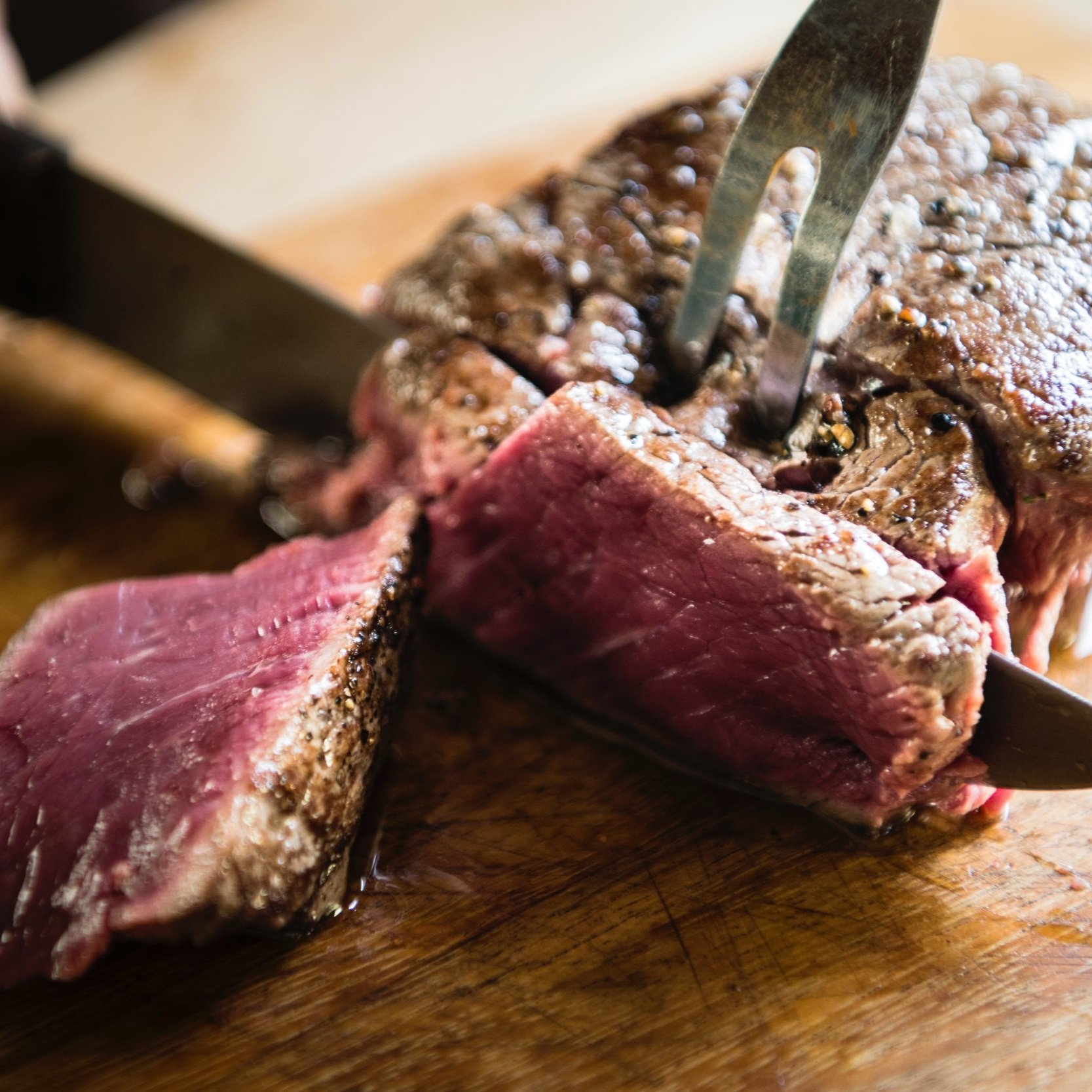 Close-up of a perfectly cooked beef roast being sliced with a knife and fork on a wooden cutting board, showcasing its juicy, pink interior and seared exterior. Ideal for culinary articles or recipes featuring beef preparation techniques.