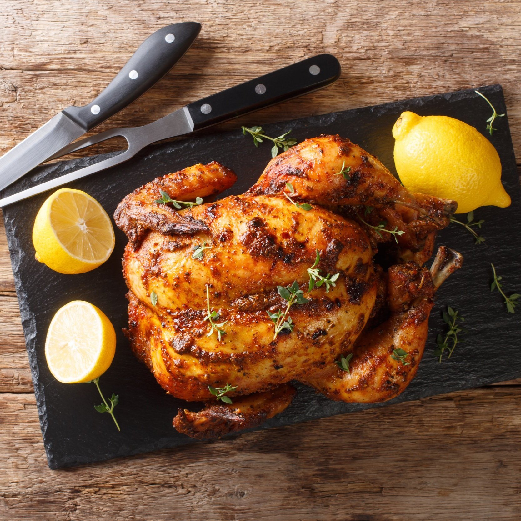 Roasted chicken seasoned with herbs and spices, garnished with fresh thyme and lemon slices, displayed on a black slate board with kitchen knives in the background.