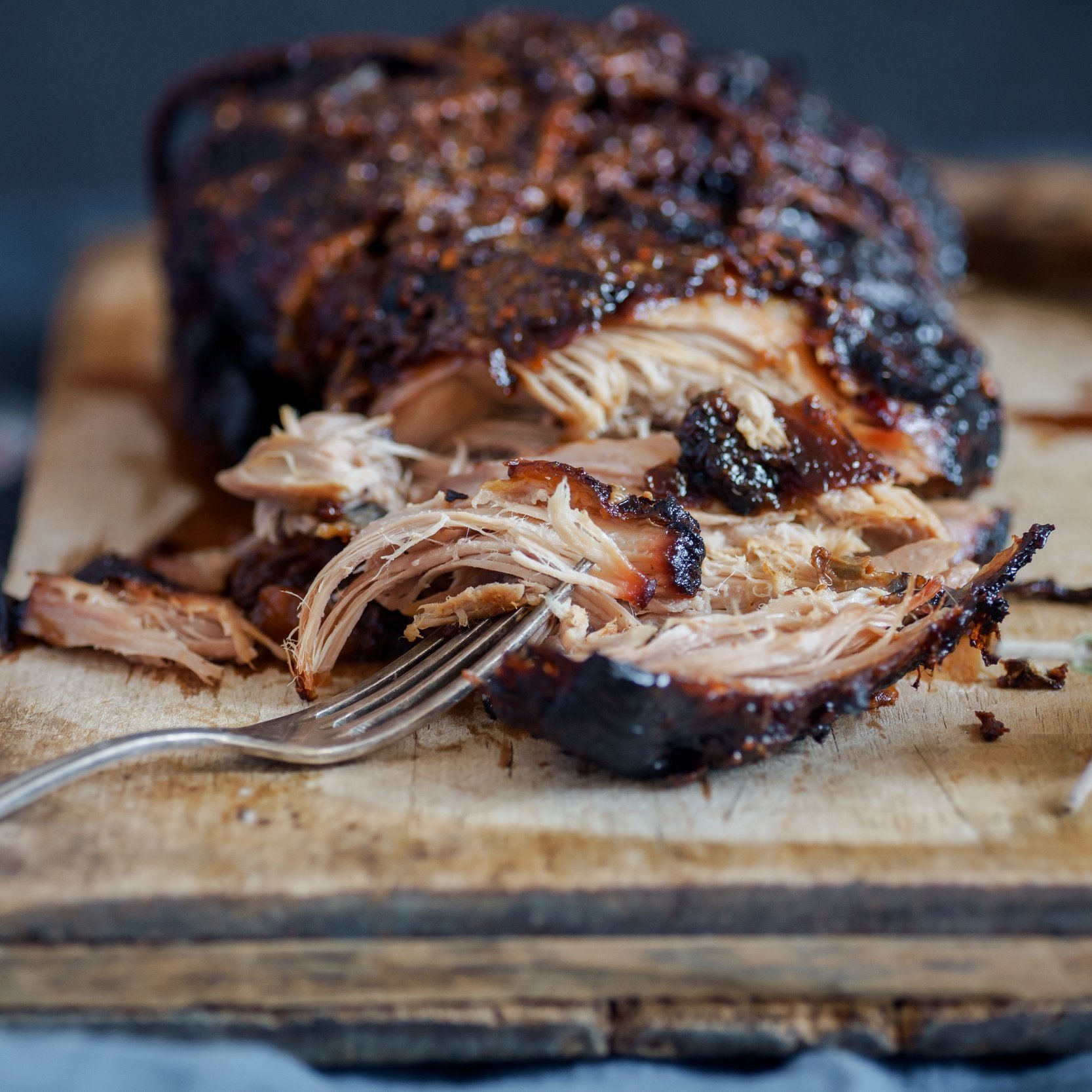 Tender pulled pork on a wooden cutting board, showcasing a fork and shredded meat, highlighting its smoky, caramelized exterior and juicy interior. Ideal for barbecue enthusiasts and recipe inspiration.